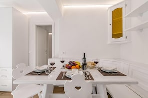 A white dining table with four chairs, a fruit bowl, wine glasses, and a bottle of wine.