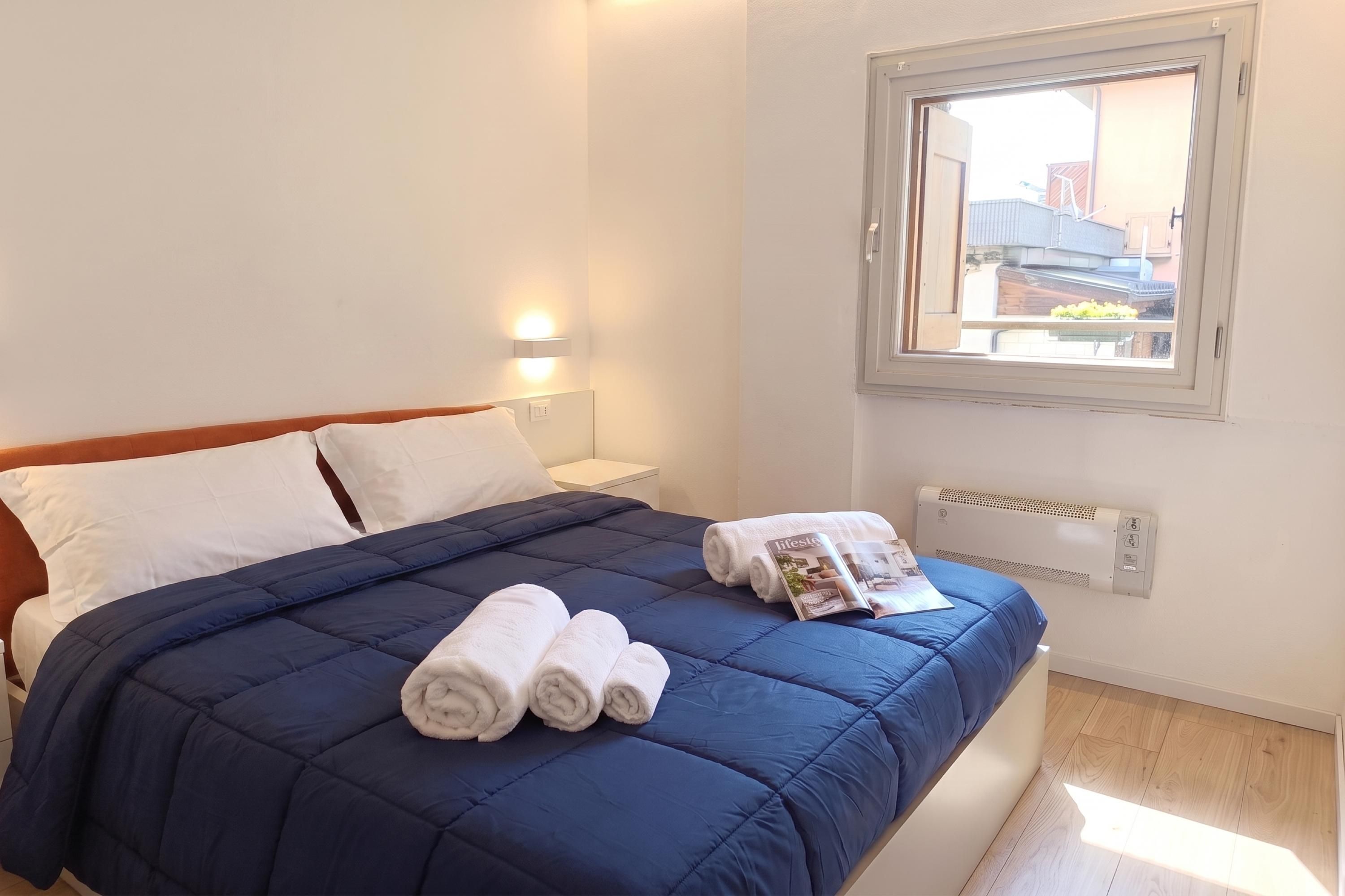 A neatly made bed with a blue duvet, white pillows, and rolled towels. There is a bedside table with a lamp, and a window with a view of buildings outside.