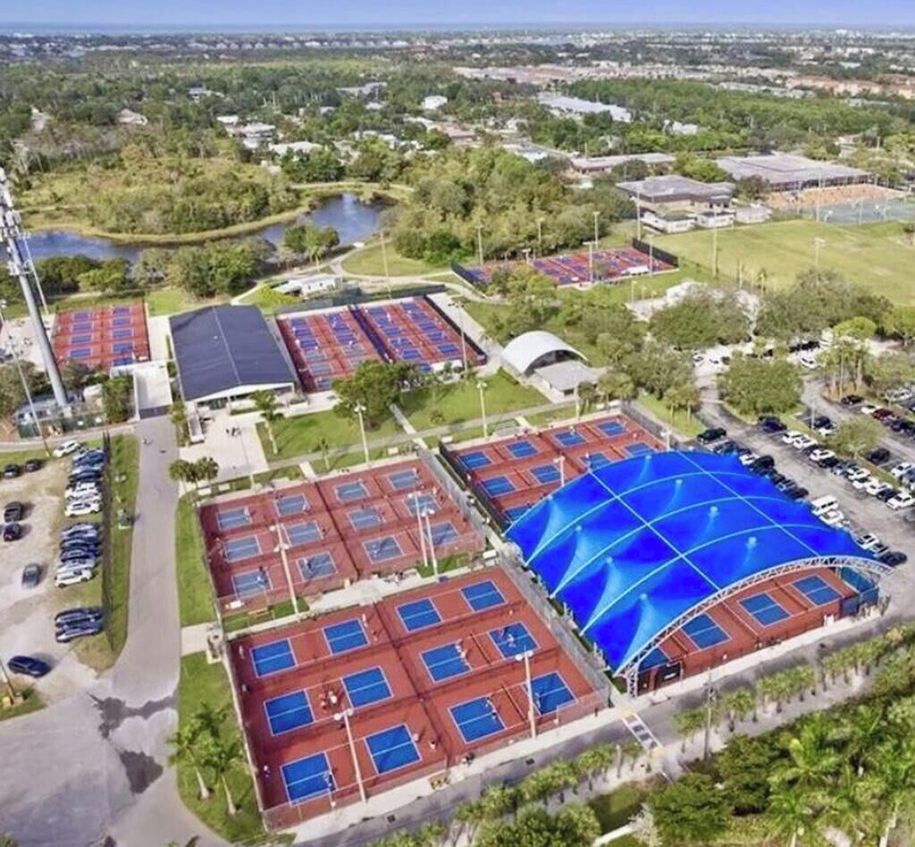 Vista aérea de un complejo deportivo con varias canchas de tenis, zonas de estacionamiento y áreas verdes circundantes.