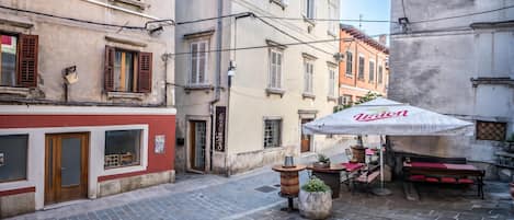 A narrow street with cobblestone pavement, traditional European buildings with shuttered windows, and a café with an umbrella.