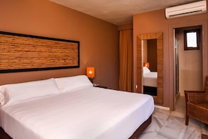 Habitación de hotel con una cama grande, una mesita de noche con lámpara, un espejo y una silla de mimbre.
