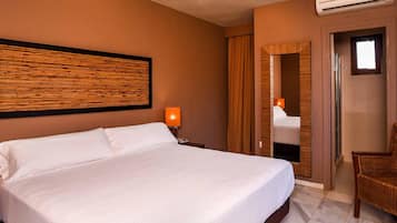 A hotel room with a large bed, a nightstand with a lamp, a mirror, and a wicker chair.
