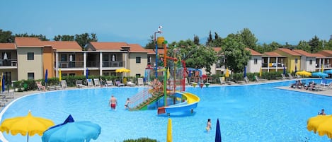 A swimming pool area with multiple water slides and umbrellas, surrounded by buildings with balconies and a clear sky.