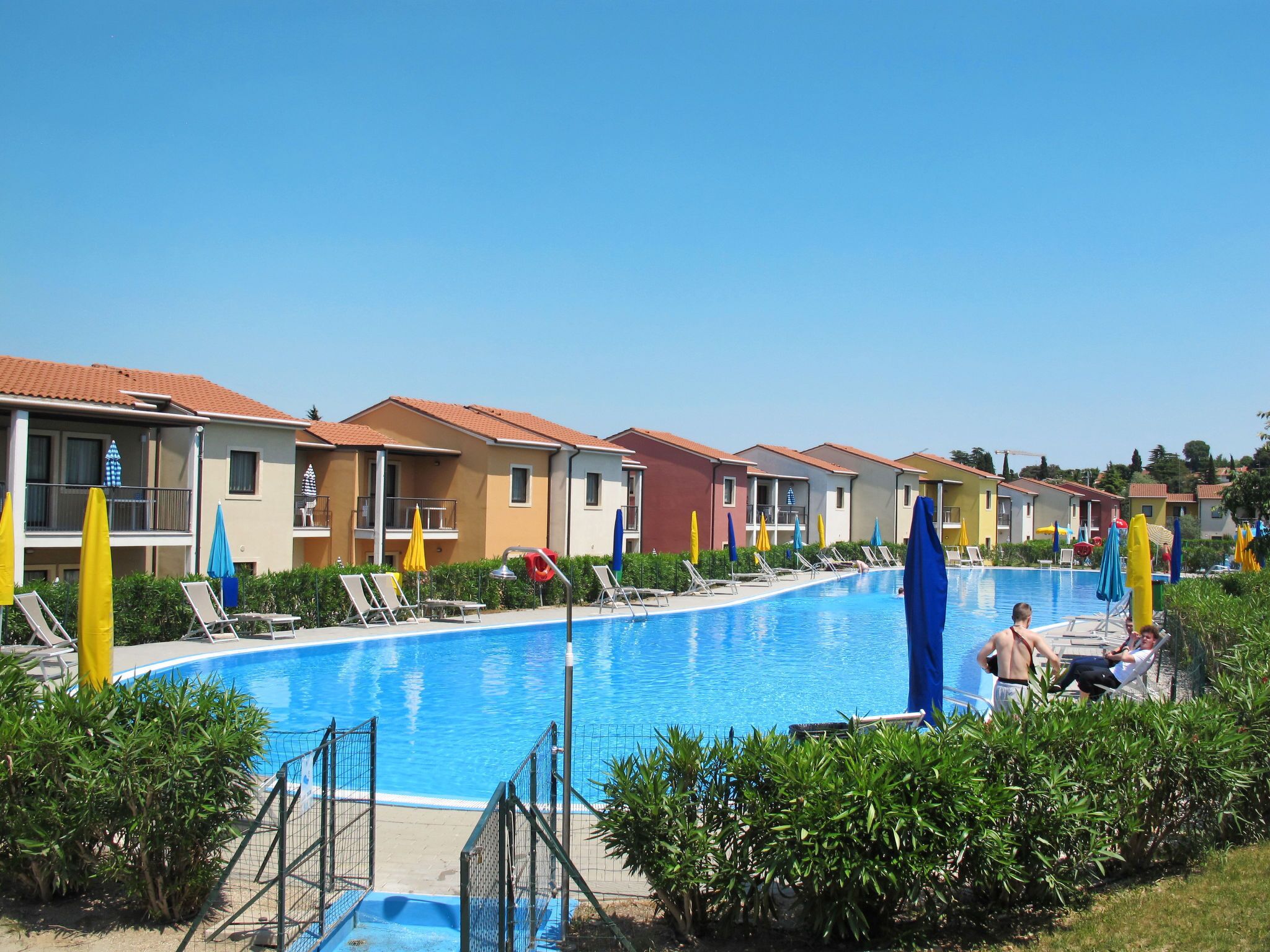 Ein Poolbereich mit mehreren Liegestühlen und Sonnenschirmen, umgeben von Gebäuden mit Balkonen.