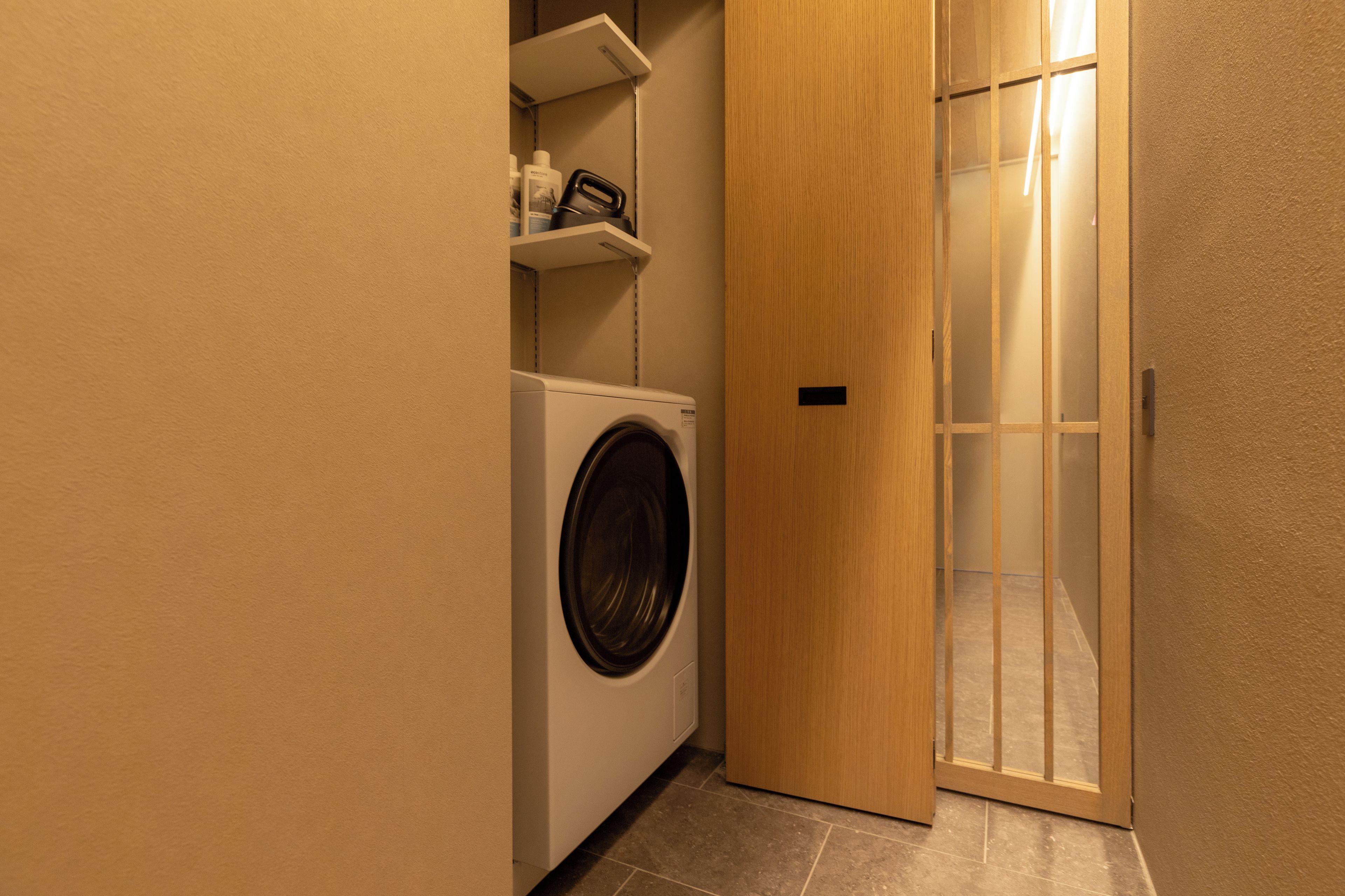 A laundry room with a washing machine, a shelf with toiletries, and a wooden door leading to another room.