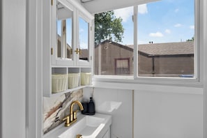 Ein Badezimmer mit weißem Waschtisch, goldenem Wasserhahn und einem Fenster mit Blick auf ein Haus.