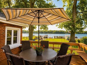 Eine Terrasse mit einem runden Tisch und Stühlen, einem gestreiften Sonnenschirm und Blick auf einen See.