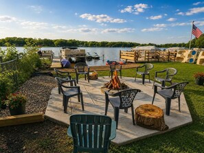 An outdoor patio with chairs and a fire pit, overlooking a lake with boats and a dock. - Lakeside Vista | 4 Seasons Waterfront Resort (Elkhart)