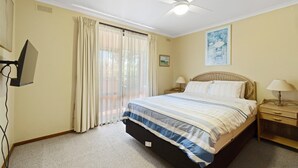 Ein Schlafzimmer mit einem Bett, zwei Nachttischen, einem Deckenventilator, einem Fenster mit Vorhängen und einem an der Wand befestigten Fernseher.
