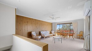 A modern living room with a wooden accent wall, a ceiling fan, a dining area with wicker furniture, and a large window with curtains.