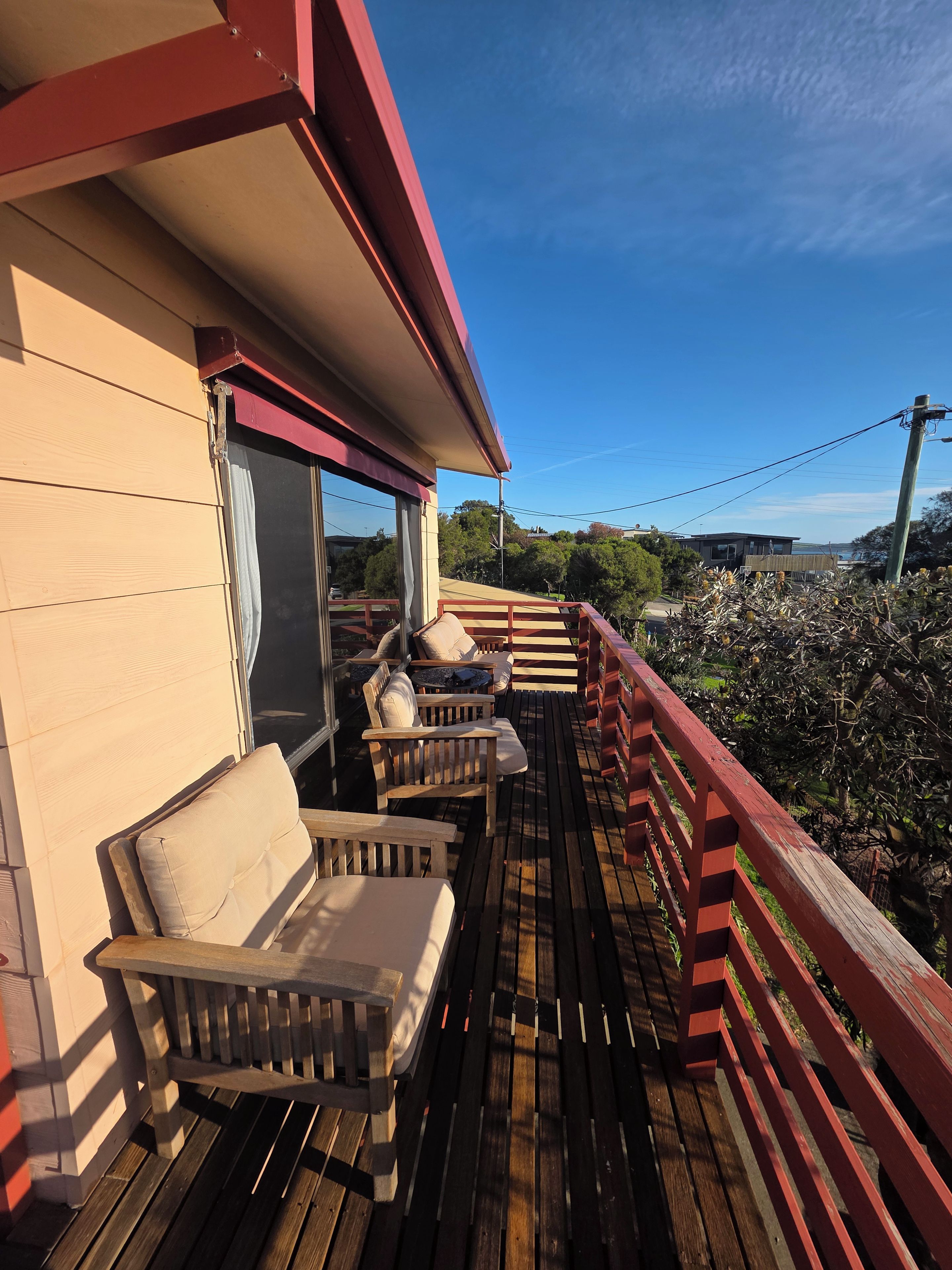 A wooden deck with outdoor furniture, a red railing, and a view of trees and a building in the background.