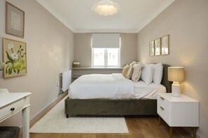 Une chambre à coucher avec un lit, des tables de chevet, un radiateur, une fenêtre et des œuvres d’art accrochées aux murs.