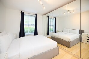 A modern bedroom with a large bed, a bedside table, a mirror, and a window with curtains. - Meet Me in Willesden Green (London)