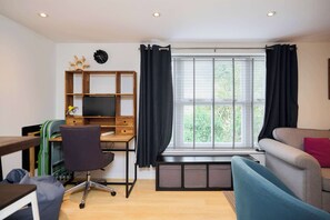 A modern living room with a wooden desk, a comfortable sofa, and a large window with curtains. - Meet Me in Willesden Green (London)