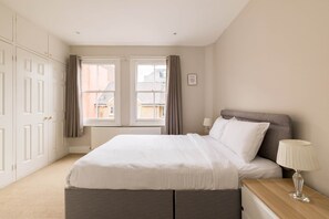 A bedroom with a bed, bedside table, lamp, and a view of buildings outside the window. - Brick & Beam on Tabard (London)