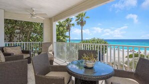 Un espacio de estar al aire libre techado con mobiliario de mimbre y una mesa, con vistas a una playa con palmeras y el océano.