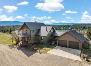 A two-story house with a garage, a covered porch, and a view of a forested area with mountains in the background. - 4-bedroom farmhouse in tranquil Ovando with AC comfort (Ovando)