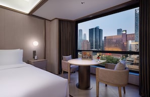 Habitación de hotel con cama, una mesa redonda con sillas, vista a un paisaje urbano y una ventana con cortinas.