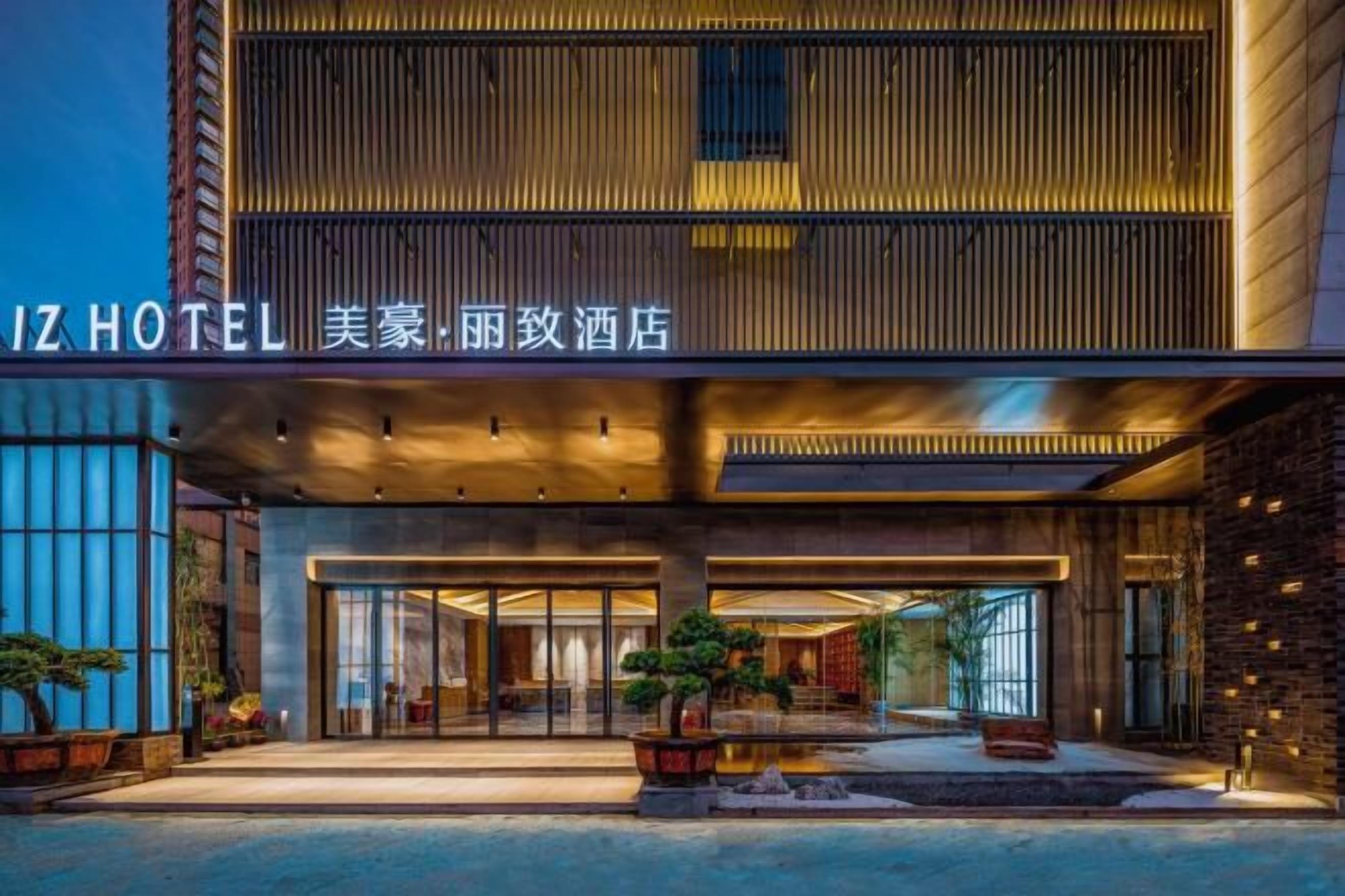 A hotel entrance with a modern design, featuring a large signboard, a covered entrance area, and potted plants.