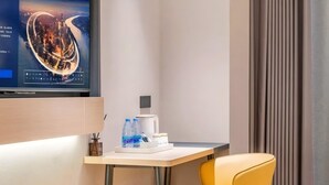 Hotellihuone, jossa on televisio, jolta näkyy kaupunkinäkymä, pieni pöytä, jossa on kahvinkeitin ja vesipulloja, sekä keltainen tuoli.