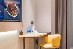 Habitación de hotel con televisor mostrando un paisaje urbano, una mesita con cafetera y botellas de agua, y una silla amarilla.