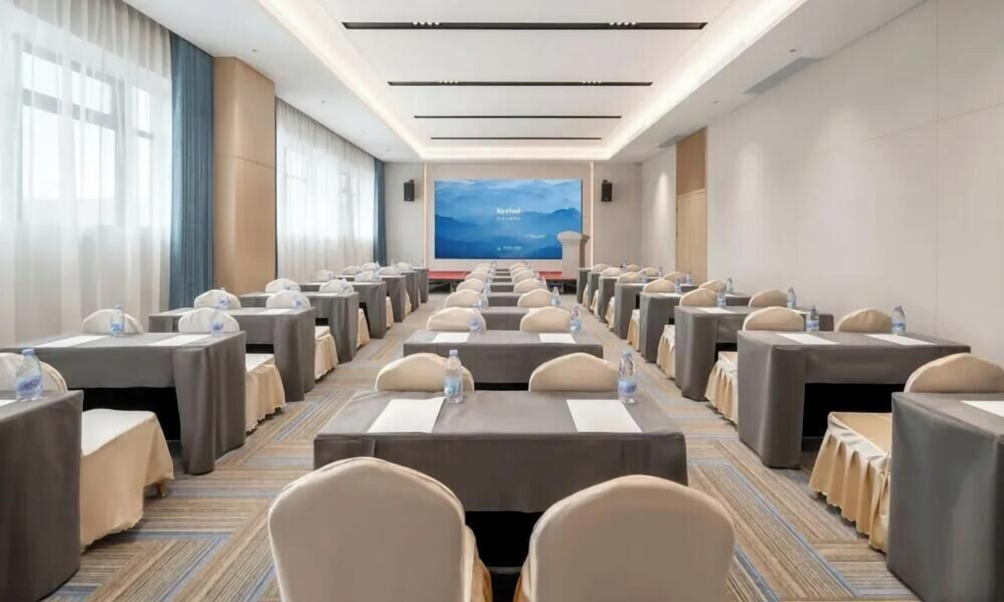 A conference room with rows of tables and chairs, a large screen at the front, and a carpeted floor.