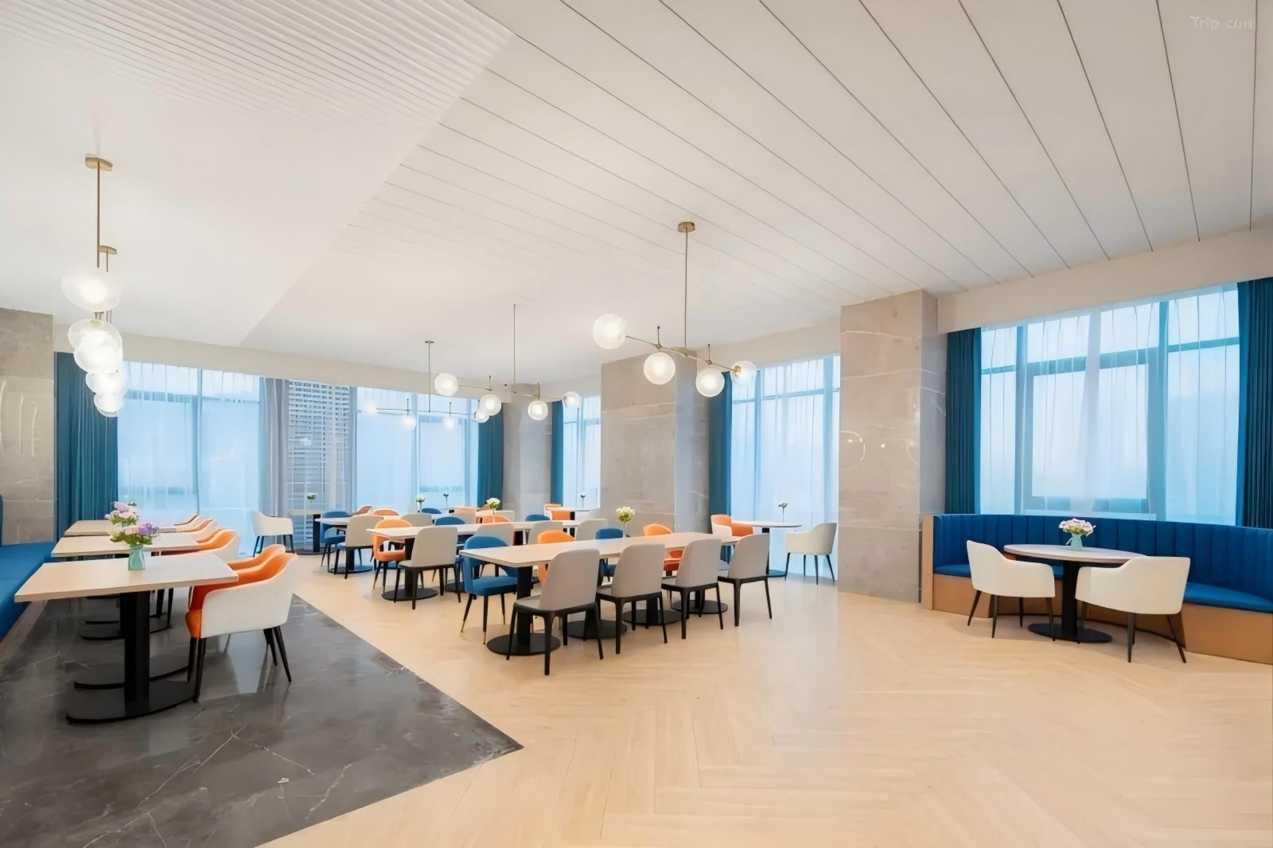 A modern dining area with multiple tables and chairs, large windows, and a high ceiling with hanging lights.