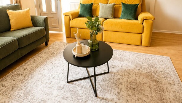 Un salon avec un canapé jaune, un canapé vert, une table basse ronde et un tapis à motifs.