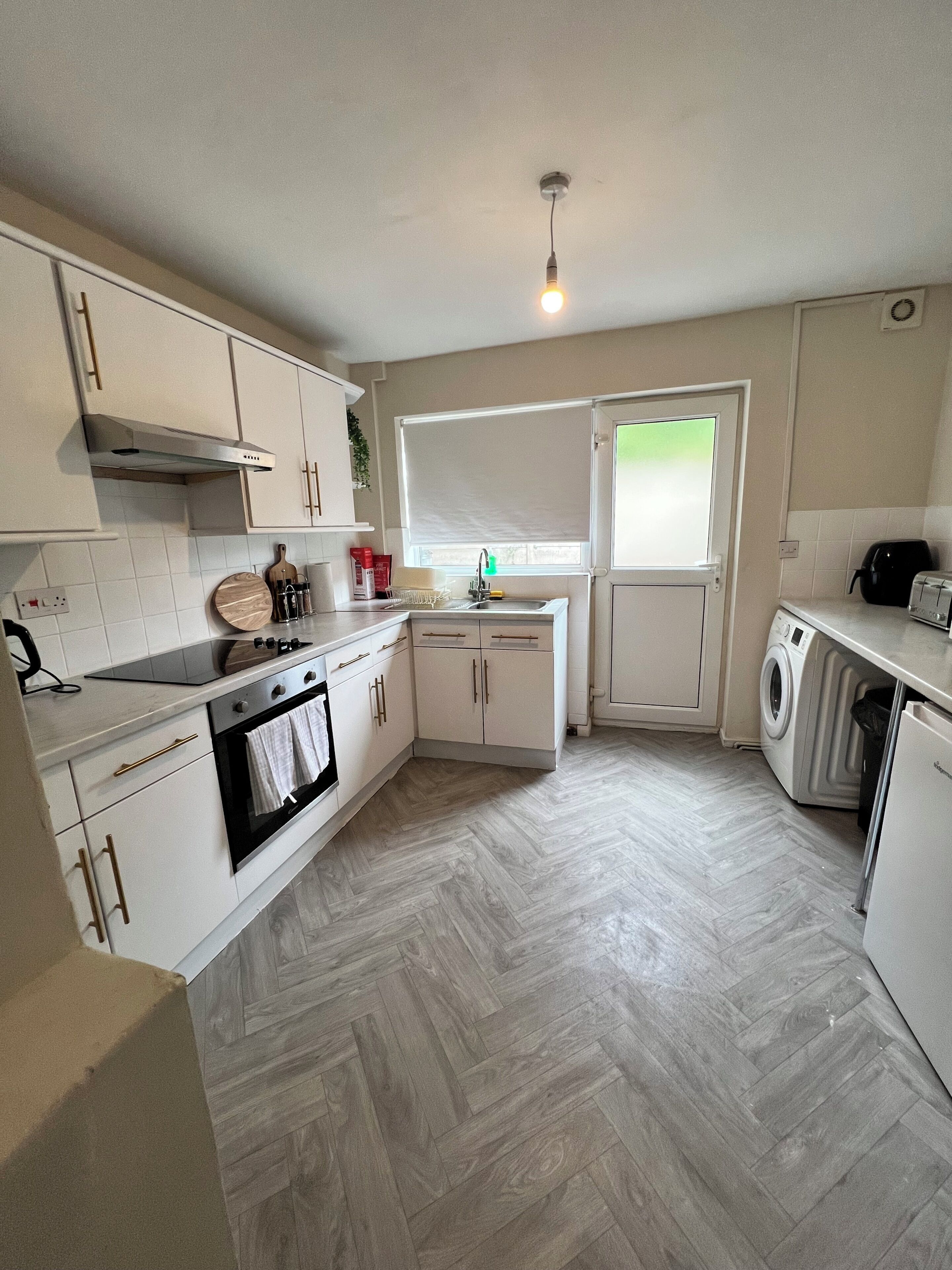 A modern kitchen with white cabinetry, a built-in oven, a washing machine, and a window with blinds.