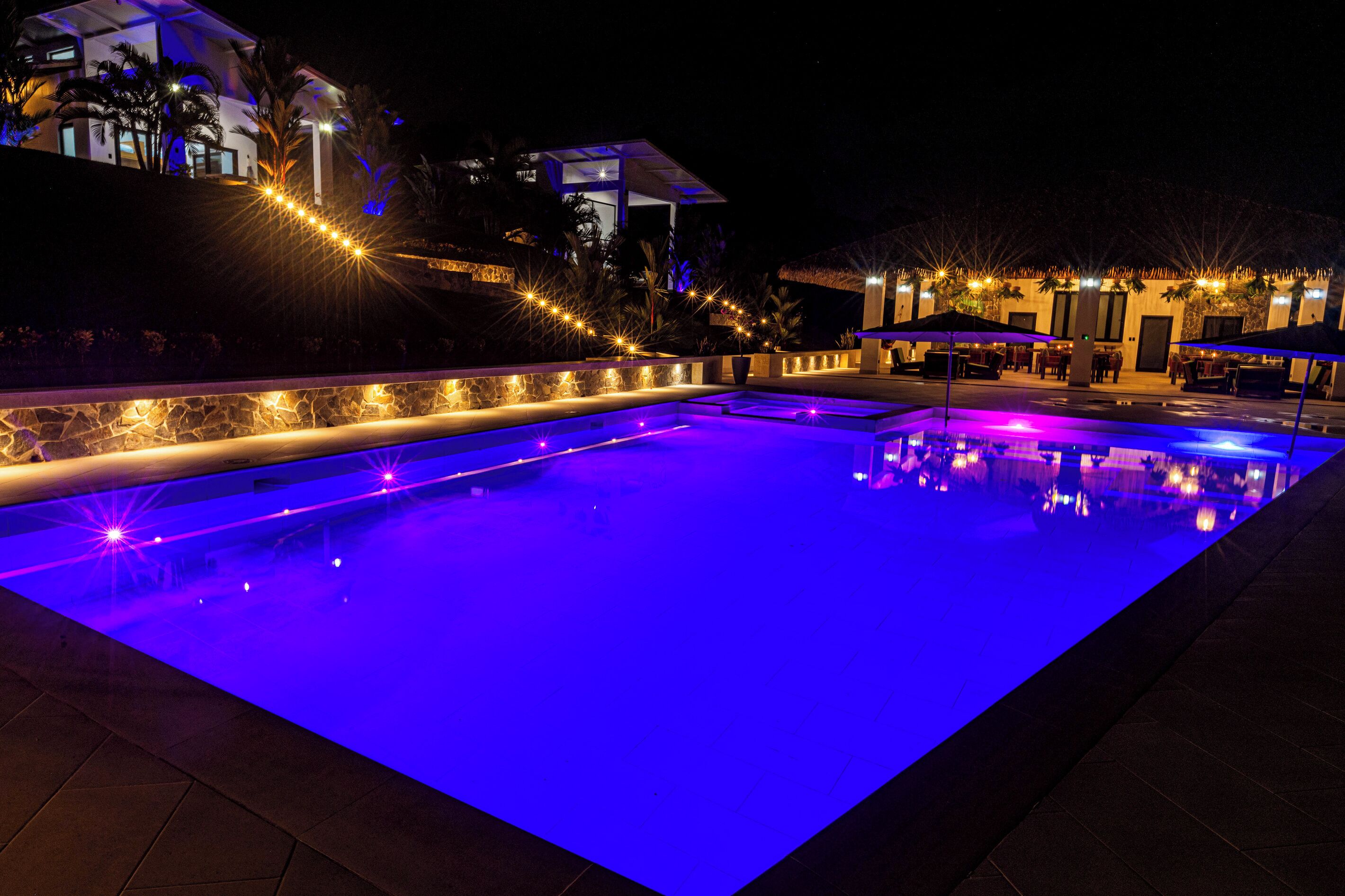 A swimming pool with a stone wall and a covered area with tables and umbrellas.