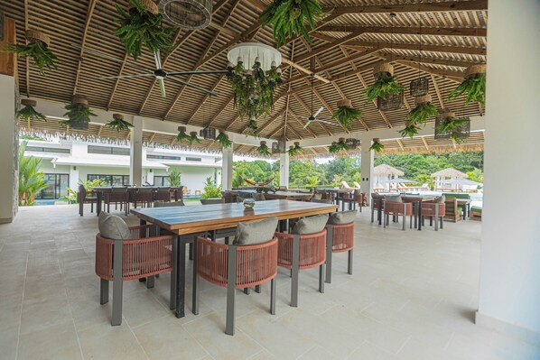 An outdoor dining area with wooden tables and chairs, hanging plants, and a thatched roof.