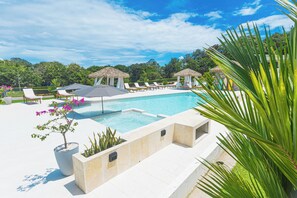 A swimming pool with lounge chairs and thatched umbrellas, surrounded by greenery and a clear sky.