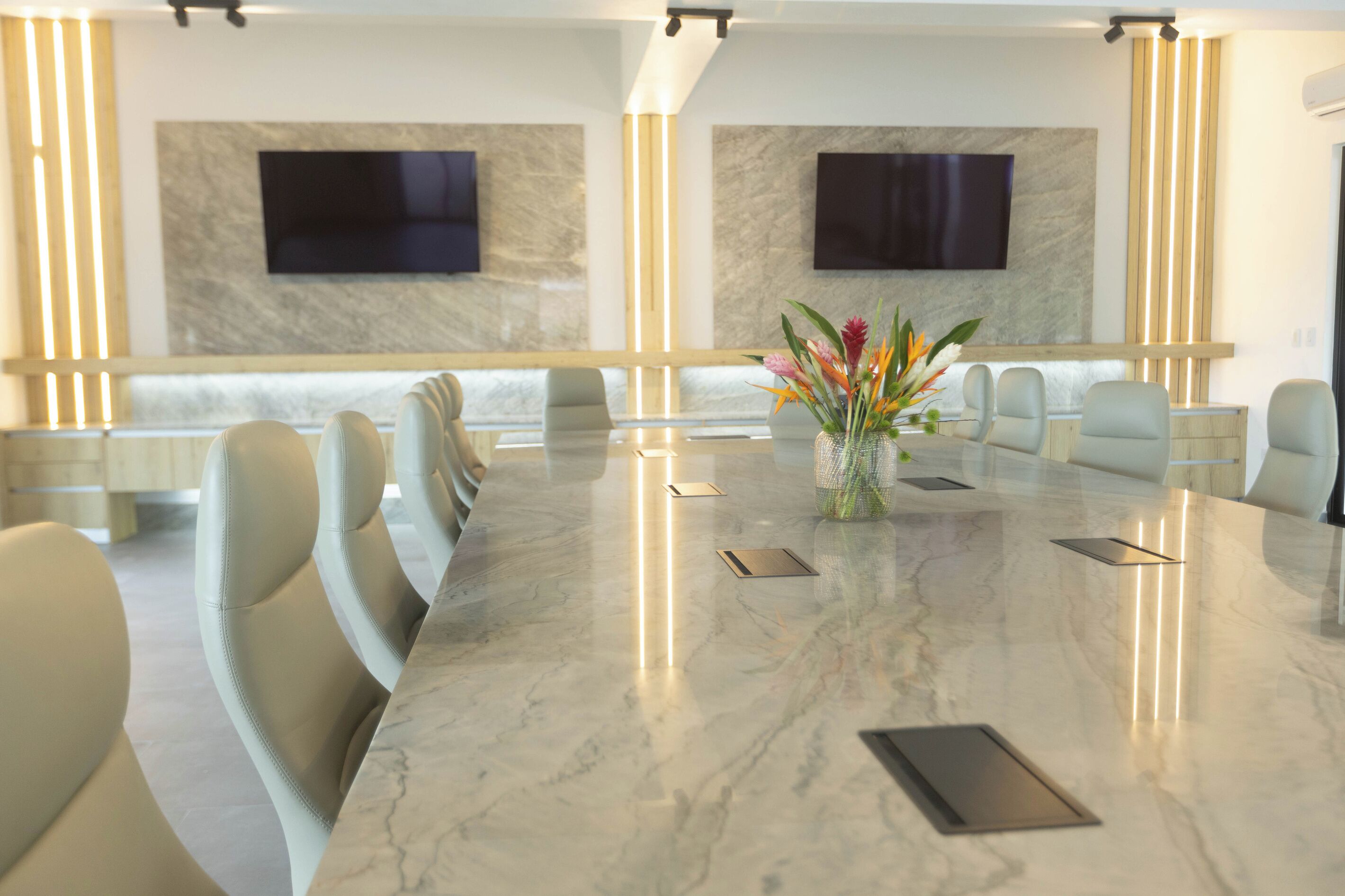 Une salle de conférence avec une table en marbre, des chaises de couleur beige, un vase rempli de fleurs et deux téléviseurs fixés au mur.