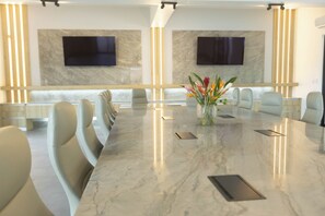 A conference room with a marble table, beige chairs, a vase with flowers, and two mounted TVs on the wall.