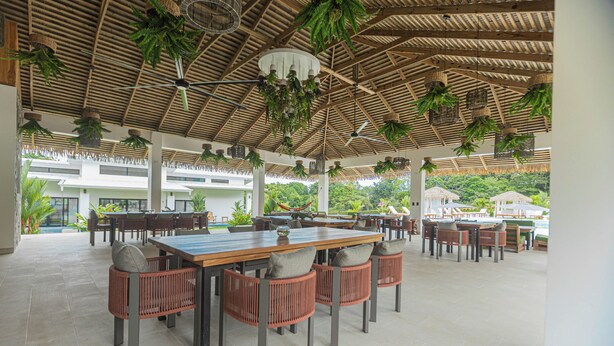 An outdoor dining area with wooden tables and chairs, hanging plants, and a thatched roof.
