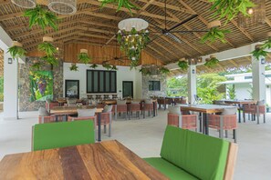 An open-plan dining area with wooden tables and chairs, a stone wall with artwork, and hanging plants.