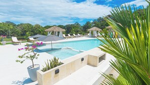 A swimming pool with lounge chairs and thatched umbrellas, surrounded by greenery and a clear sky.