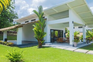 Ein modernes Haus mit überdachter Terrasse, Rasenfläche und einer Palme.