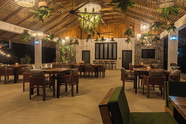 A spacious indoor area with multiple wooden tables and chairs, hanging plants, and a high ceiling with exposed beams.