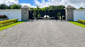 Une entrée fermée par un portail, une allée gravillonnée, des murs en pierre et un ciel d’un bleu éclatant.