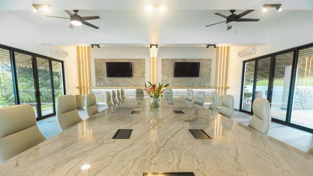 Une salle de conférence dotée d’une table en marbre, de chaises beiges, de ventilateurs de plafond et de grandes fenêtres.