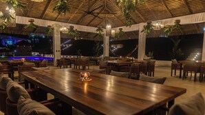 Un espace extérieur aménagé pour les repas, avec des tables et des chaises en bois, des plantes suspendues et un toit de chaume.