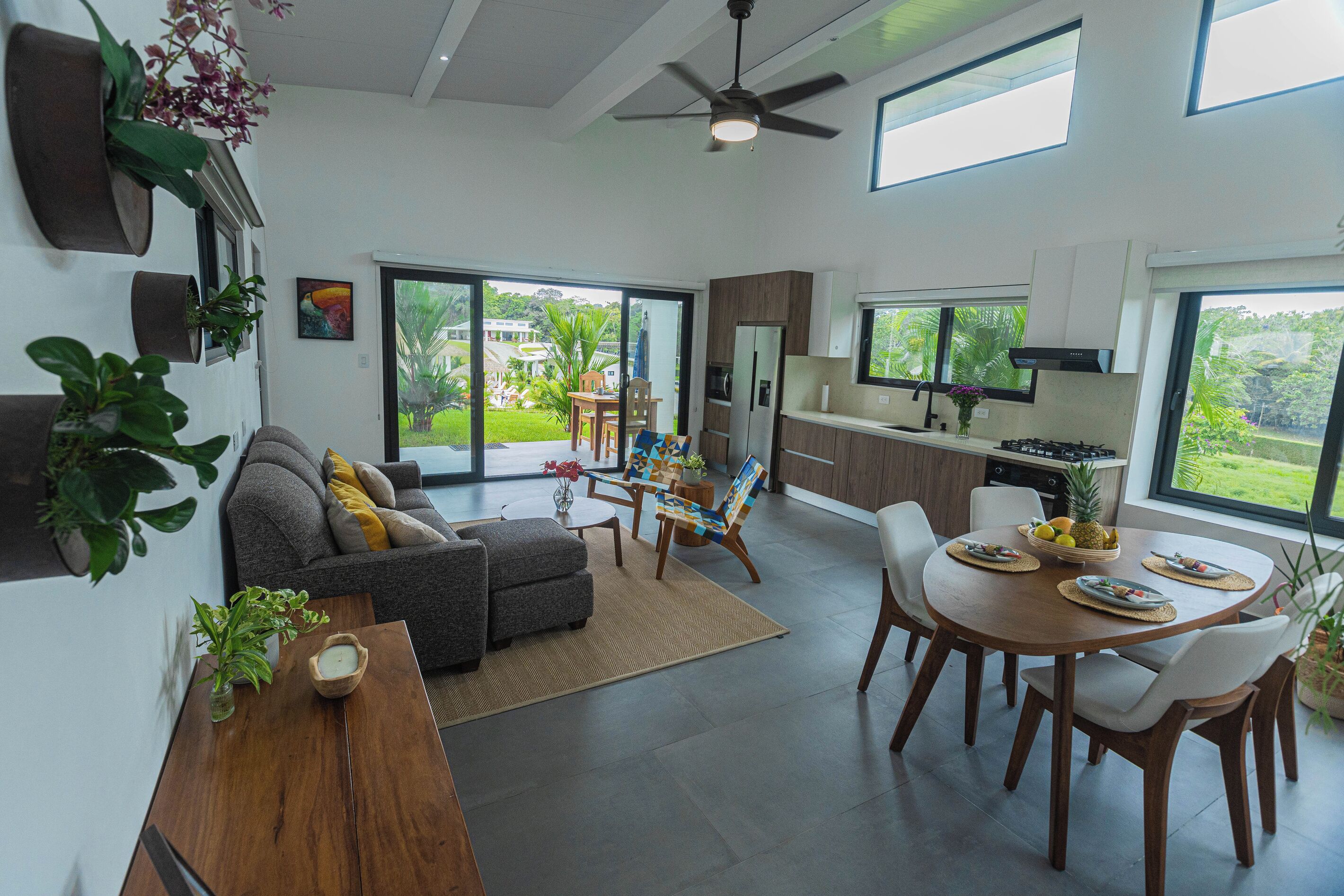 Una sala moderna con sofá, una mesa de comedor con sillas, una zona de cocina con estufa y ventanas amplias que ofrecen vistas al exterior.