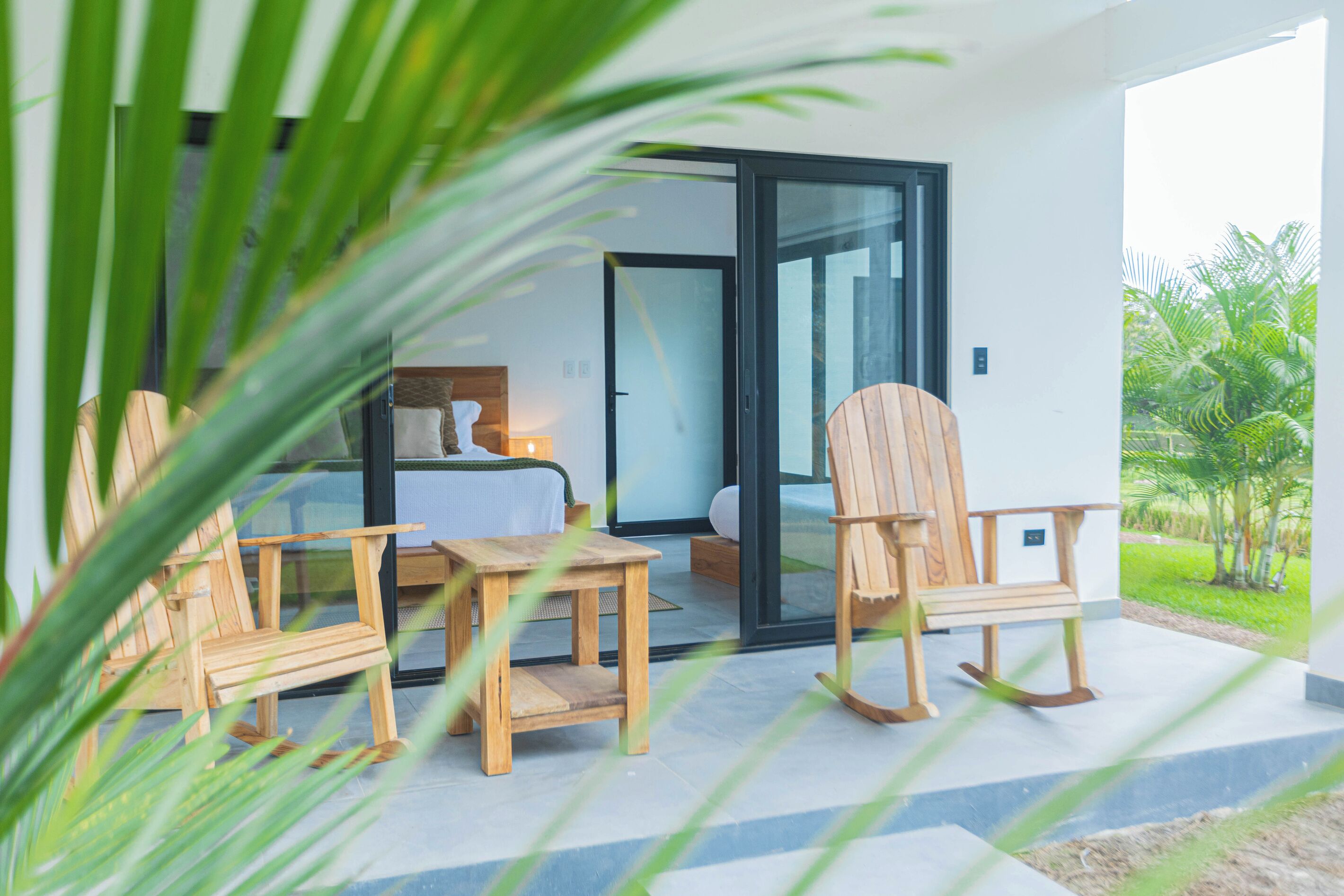A room with a bed, a wooden chair, a small table, and a sliding glass door leading to a balcony with a view of greenery.