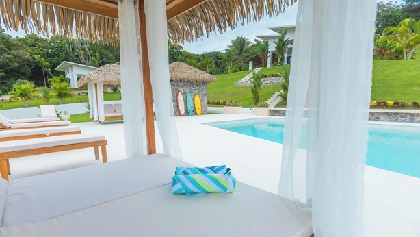 Un espace aménagé au bord de la piscine, avec des chaises longues, un lit et une structure dotée d’un toit de chaume.