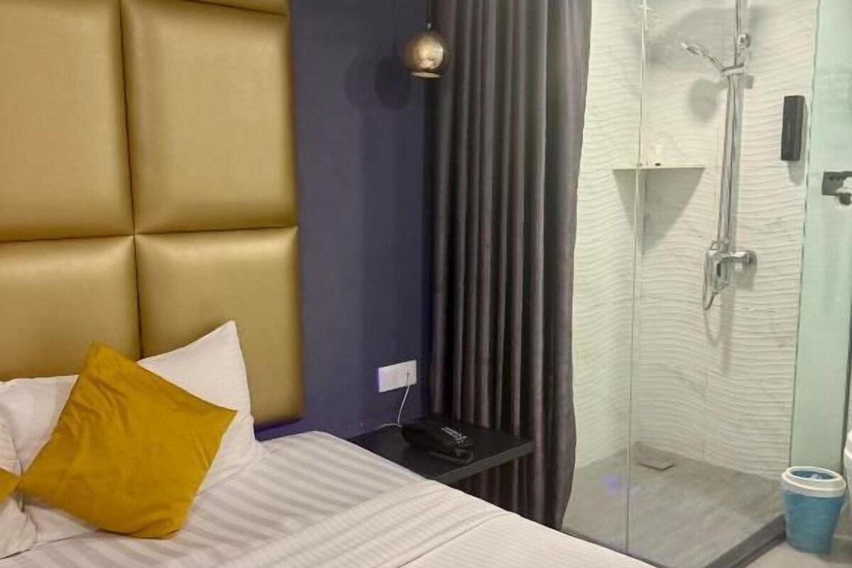 A hotel room with a bed, a glass shower enclosure, and a bedside table with a lamp.