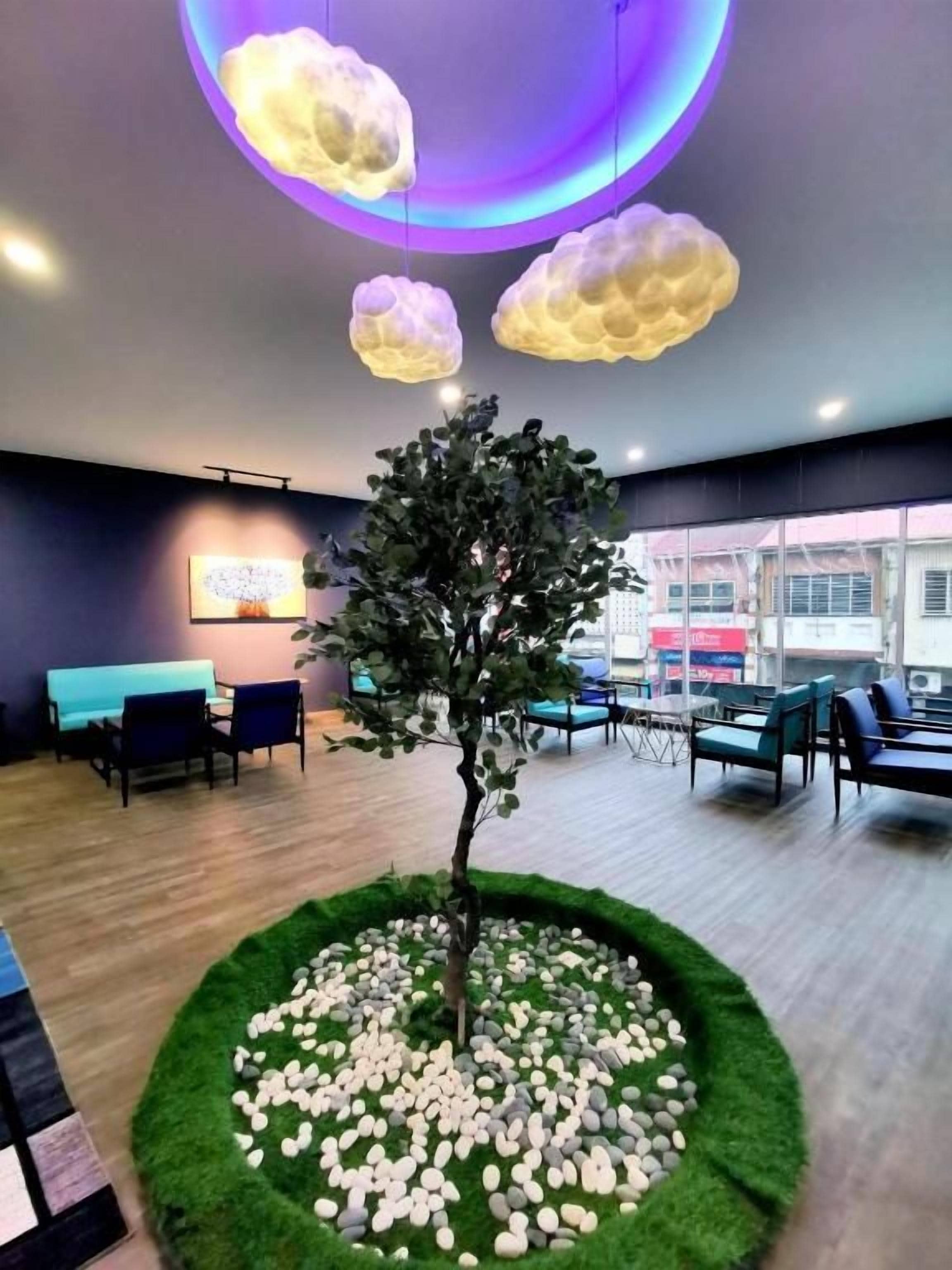 Un interior moderno con un árbol central rodeado por una disposición circular de piedras, una zona de estar con sillas e iluminación en el techo con forma de nubes.