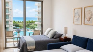 A hotel room with a bed, a sofa, a small table, and a lamp. There is a window with a view of a pool and a building. - Suite Apartment | Ocean View | Infinity Pool | Rooftop | Jacuzzi (Santa Marta)