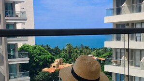 A person reading a book on a balcony overlooking a pool and a view of a coastal area with buildings and palm trees. - Suite Apartment | Ocean View | Infinity Pool | Rooftop | Jacuzzi (Santa Marta)