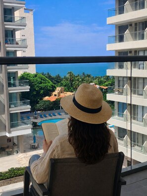 Eine Person liest ein Buch auf einem Balkon mit Blick auf einen Pool und die Küste, mit Gebäuden und Palmen.